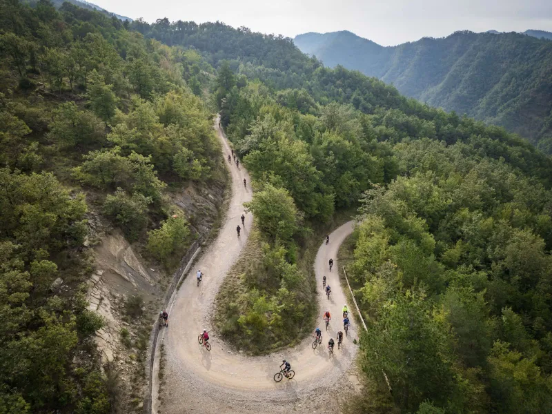 Una strada bianca che si divide in due nel bosco delle Foreste Casentinesi, con i ciclisti in fila, vista dall'alto