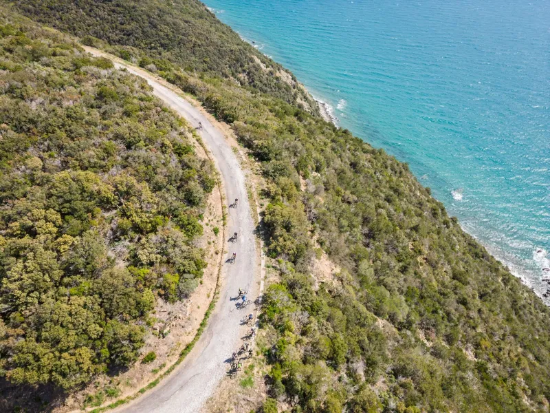 Una strada bianca che corre tra la macchia mediterranea e il mare turchese, con alcuni ciclisti, vista dal drone
