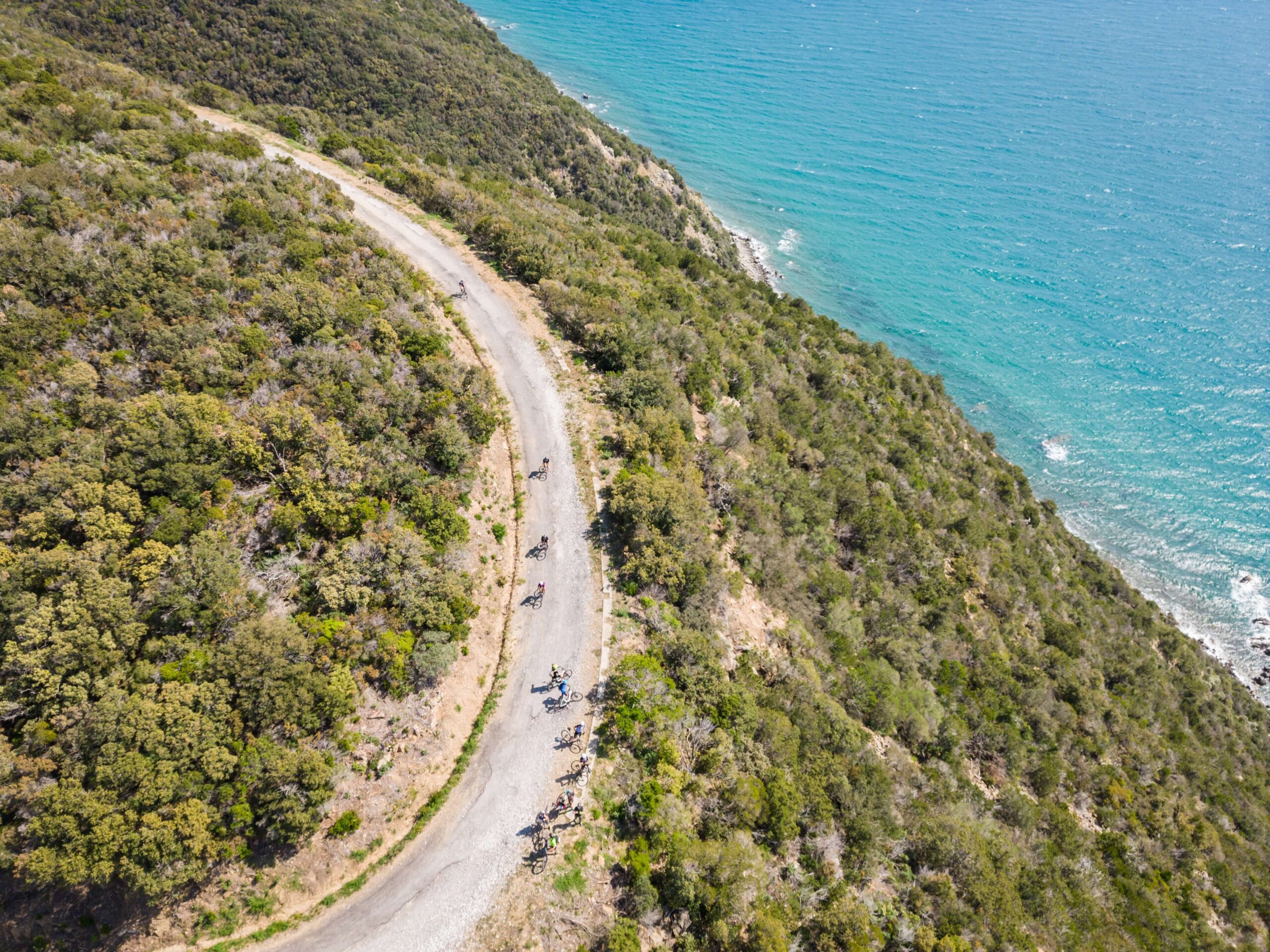 Vista aerea di una strada costiera sterrata che si snoda lungo una collina verdeggiante affacciata su un mare blu cristallino. Un gruppo di ciclisti in bicicletta percorre la strada, immersi in uno scenario naturale mozzafiato.