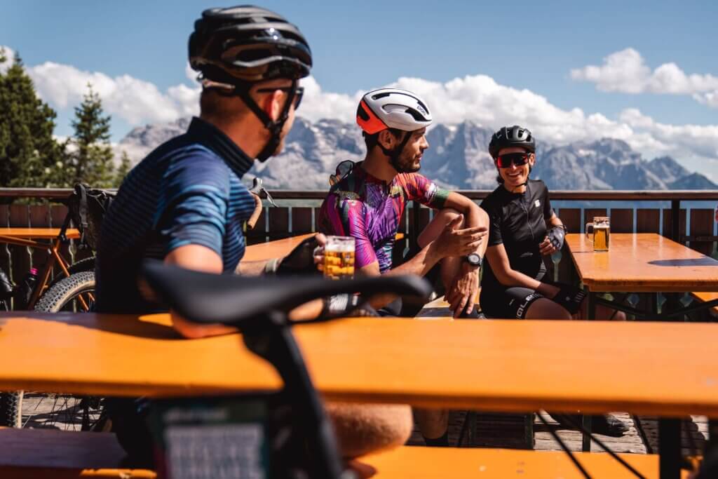 Un gruppo di ciclisti gravel si rilassa su una terrazza panoramica in montagna, sorseggiando birra e chiacchierando dopo una pedalata. Sullo sfondo, un maestoso paesaggio alpino con montagne e cielo azzurro arricchisce l’atmosfera conviviale.